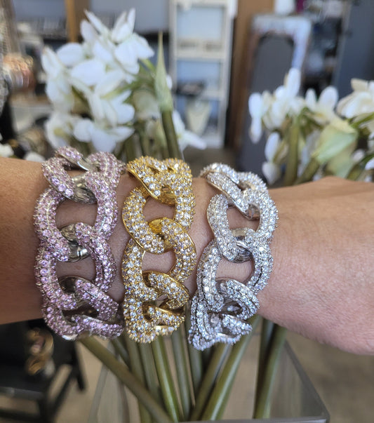 A forearm displays three Curved Link link CZ Bracelet Zirconite couture.582B1338 bracelets by DiamondVeneer Fashion in pink, gold, and silver, all sparkling with rhinestones. White flowers and blurred shelves are visible in the indoor background.