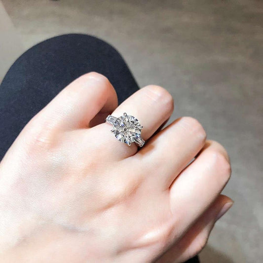 A hand wearing the DiamondVeneer Fashion Round Solitaire Baguette Diamond Veneer Cubic Zirconia 14K Gold Ring, featuring a large round clear gemstone, is displayed against a blurred background.