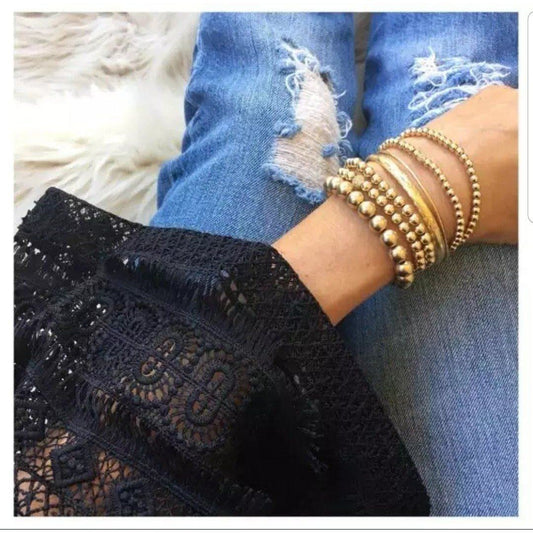 A person wearing several gold bangles, including the DiamondVeneer Fashion Never Tarnish Zirconite Gold Bead Stretch Bracelet, sits cross-legged in ripped blue jeans and a black lace sleeve on a white furry surface.