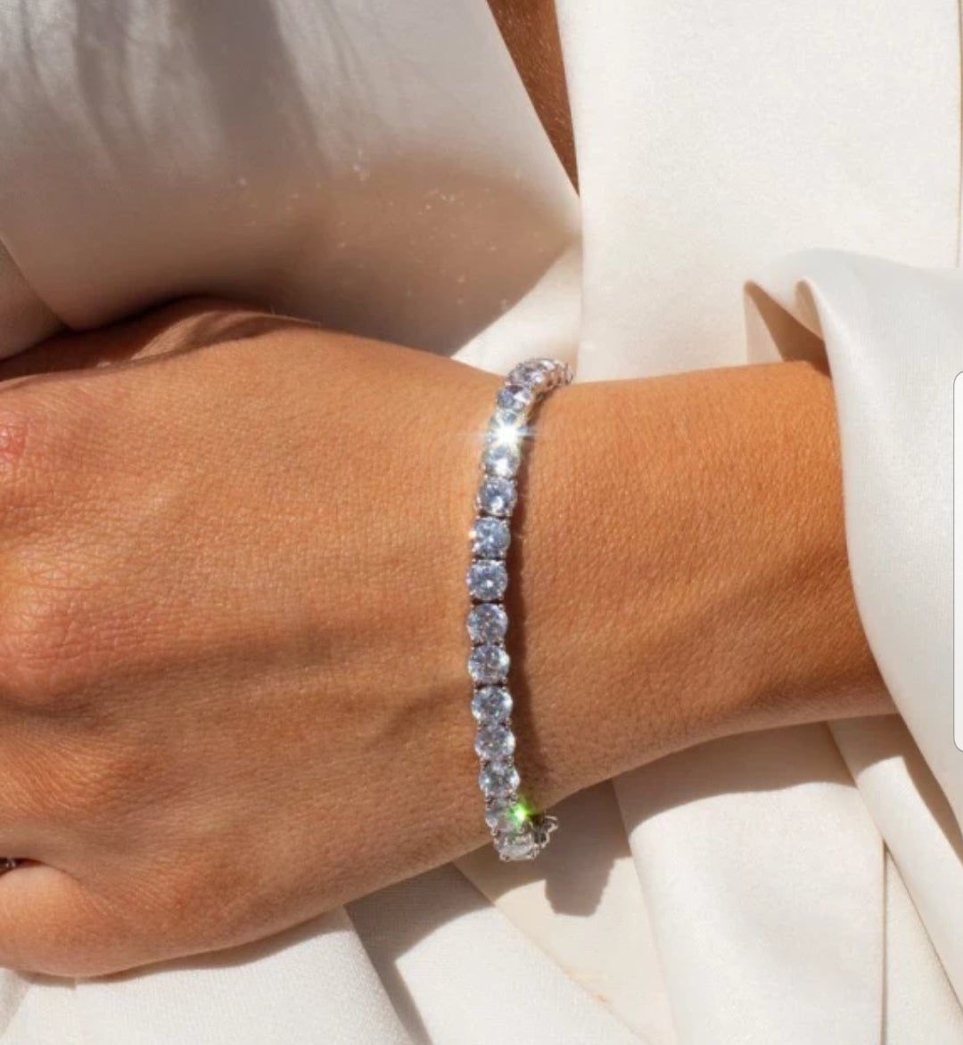 Round cubic zirconia tennis bracelet with safety chain worn on a woman's wrist against white fabric, 698Bcast.