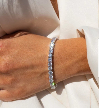Round cubic zirconia tennis bracelet with safety chain worn on a woman's wrist against white fabric, 698Bcast.